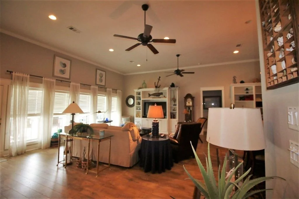 Living room with wooden floors, multiple windows with sheer curtains, ceiling fans, and various lamps. There are sofas, armchairs, and shelves with decorative items.