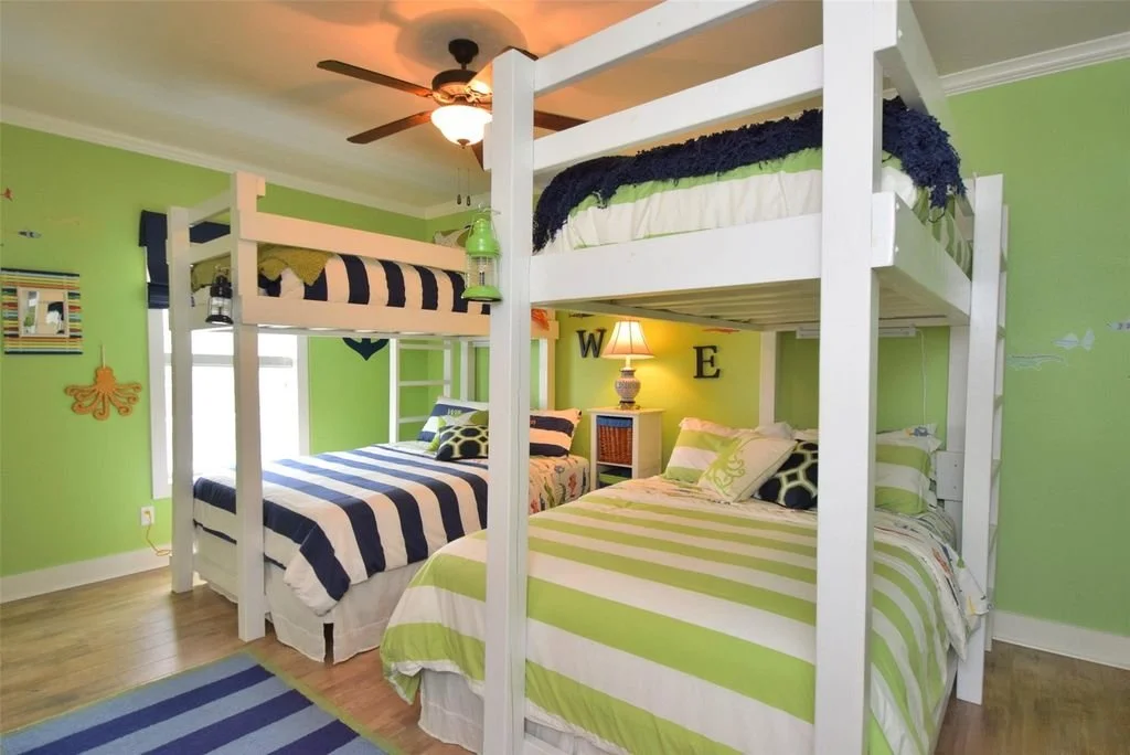 Bedroom with three beds, two with bunk bed frames, painted in green with blue and white striped bedding, and one with green and white striped bedding. A small nightstand with a lamp is between the bottom beds. Decorations include a painting, a sea cr
