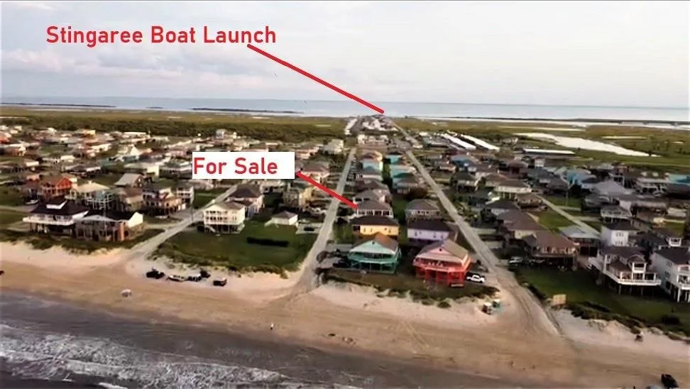 Aerial view of a beach town with houses leading to the water, featuring a sign pointing to the 'Stingaree Boat Launch' in the distance. A section of the beach has some parked cars and sandy shoreline.