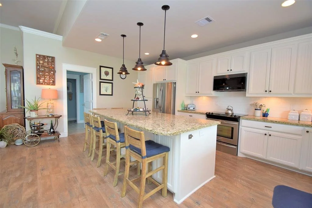 Modern kitchen with white cabinets, granite countertops, stainless steel appliances, and a central island with six wooden chairs. Pendant lights hang above the island, and there are decorative items and framed pictures on the walls.