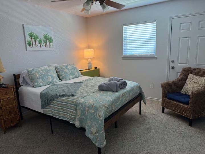 A neatly made bedroom with a bed, two pillows, a folded towel, a nightstand with a lamp, a wicker chair with a pillow, and a window with blinds.