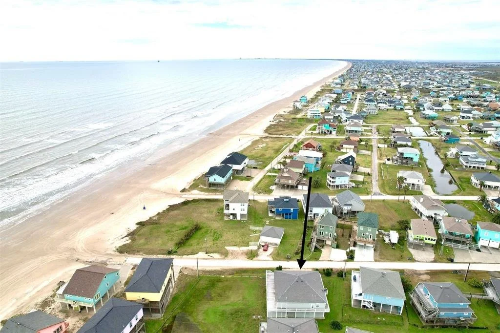 Aerial view of beachfront houses in a coastal neighborhood, with the ocean on the left and houses on the right, showing a mix of small and large homes, some with decks and porches, and a grassy vacant lot in the foreground.
