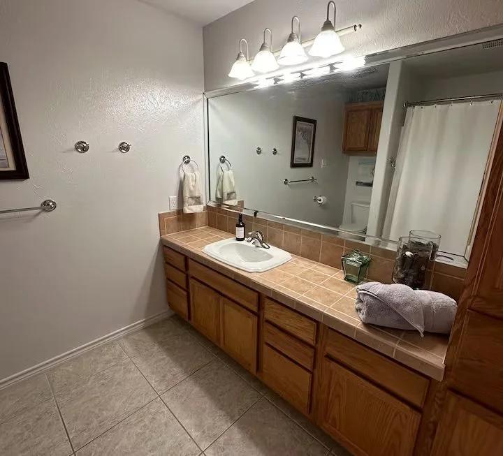 Bathroom with large mirror, wooden cabinet, beige tiled countertop with a sink, soap dispenser, towel, and toothbrush holder. There are four lights above the mirror, and a towel rack on the wall. Additional storage cabinet visible on the right.