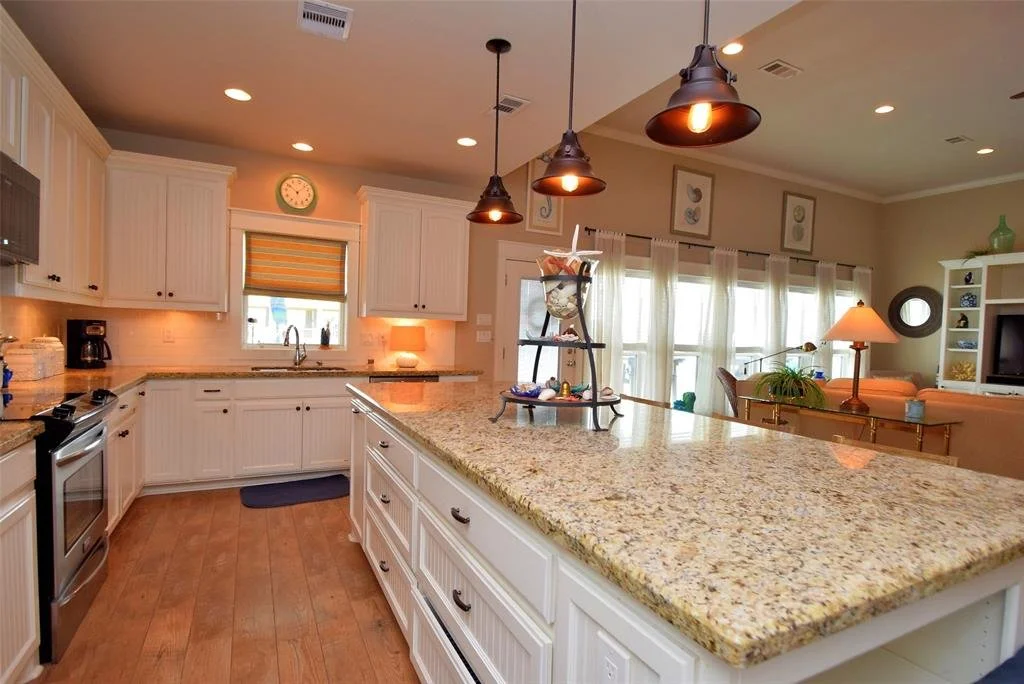 Kitchen with white cabinets, granite countertops, stainless steel oven, coffee maker, and a window above the sink. Pendant lights hang above a large center island, and the background shows a dining area with a table, chairs, and a living room with a 