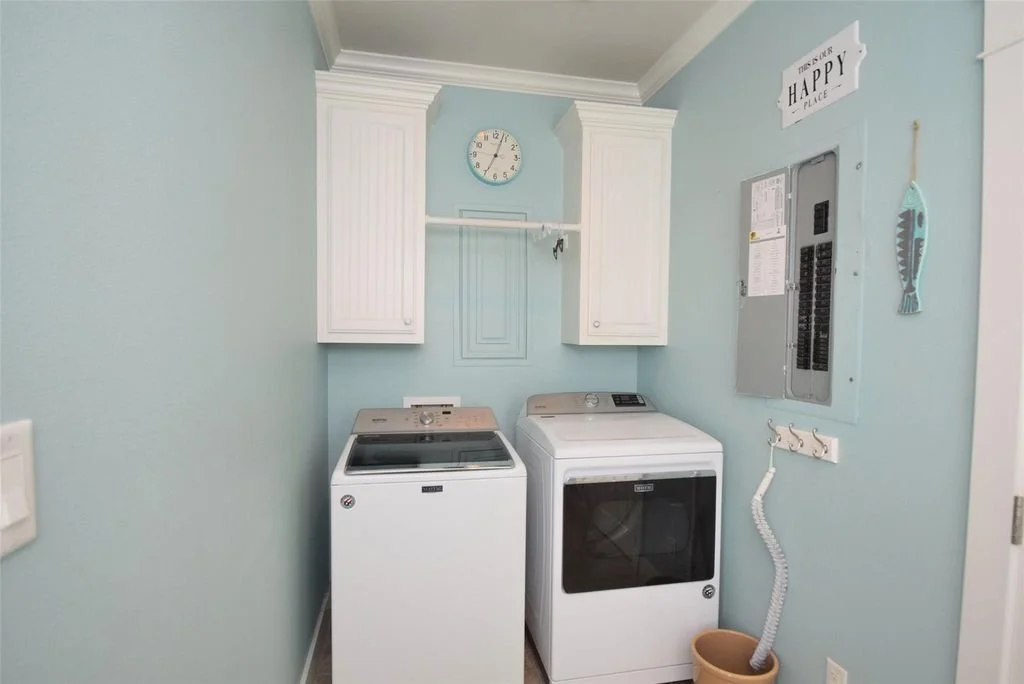 A laundry room with a white washing machine and dryer, light blue walls, white cabinets, a wall clock, a sign that reads 'This is my happy place', and a fish wall decoration.