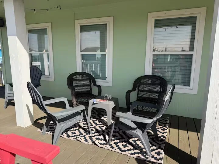 Outdoor patio with four black and white chairs on a black and white patterned rug, three windows with blinds on a light green wall, and string lights overhead.
