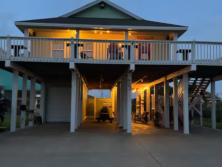 Beach house on stilts with a balcony and staircase, illuminated at dusk.