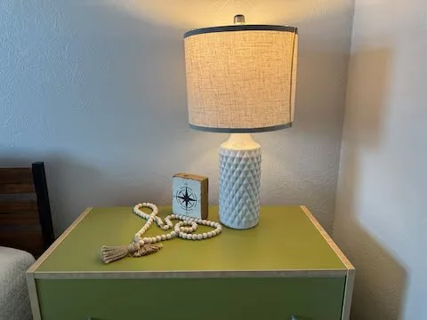 Lampshade on a textured white lamp base, placed on a green nightstand with decorative items, against a light-colored wall.