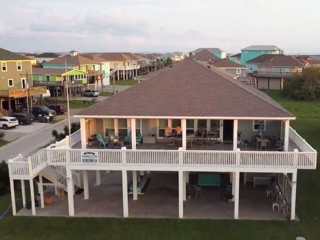 A two-story beach house with a large balcony on the second floor, furnished with chairs, tables, and outdoor decor. The house is elevated on stilts with a spacious covered area underneath.