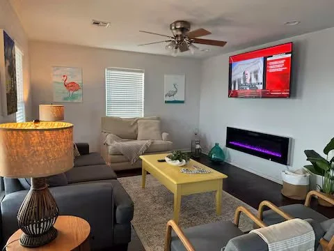 Living room with a ceiling fan, two paintings of flamingos, a window with blinds, a wall-mounted TV, a black fireplace, and various furniture including a yellow coffee table, sofas, chairs, and a lamp.