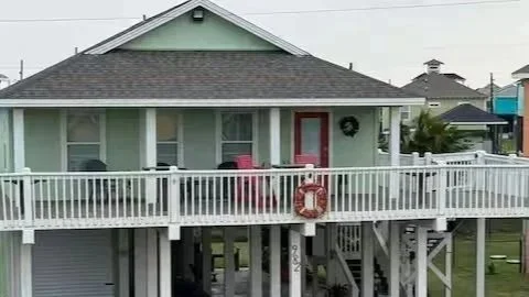 A light green house with a dark gray roof and a red door, featuring a large white porch with white railing and a red life ring hanging on it.