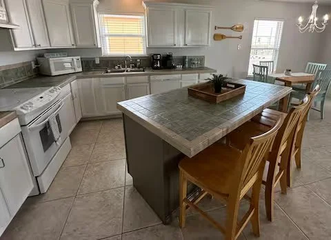 A kitchen with white cabinets, a large island with a tiled countertop, and wooden chairs around it. The kitchen has appliances such as a microwave and coffee maker, and a dining area with a table and chairs in the background.