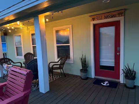 Front porch of a house with a red door, potted plants, seating area, and string lights at sunset.