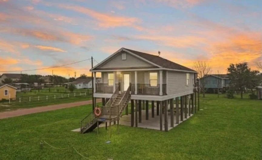 A raised house with stairs leading to a porch, situated on stilts over a grassy yard at sunset.
