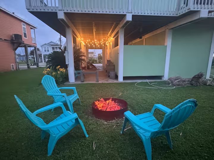 A backyard at dusk with three blue plastic chairs arranged around a round fire pit with glowing embers. There are string lights hanging under the elevated deck, and neighboring houses are visible in the background.