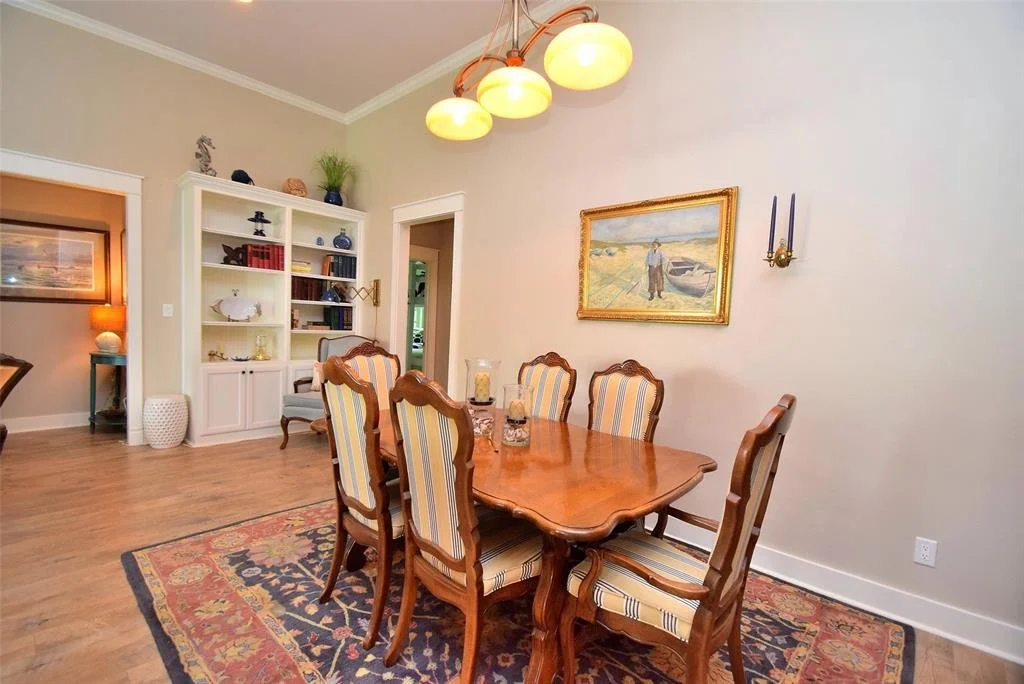 A dining room with a wooden table and six upholstered chairs with stripe-patterned fabric. There is a large area rug under the table, a painting on the wall, and a white built-in bookshelf with decorative items. Overhead, there is a brass lighting fi