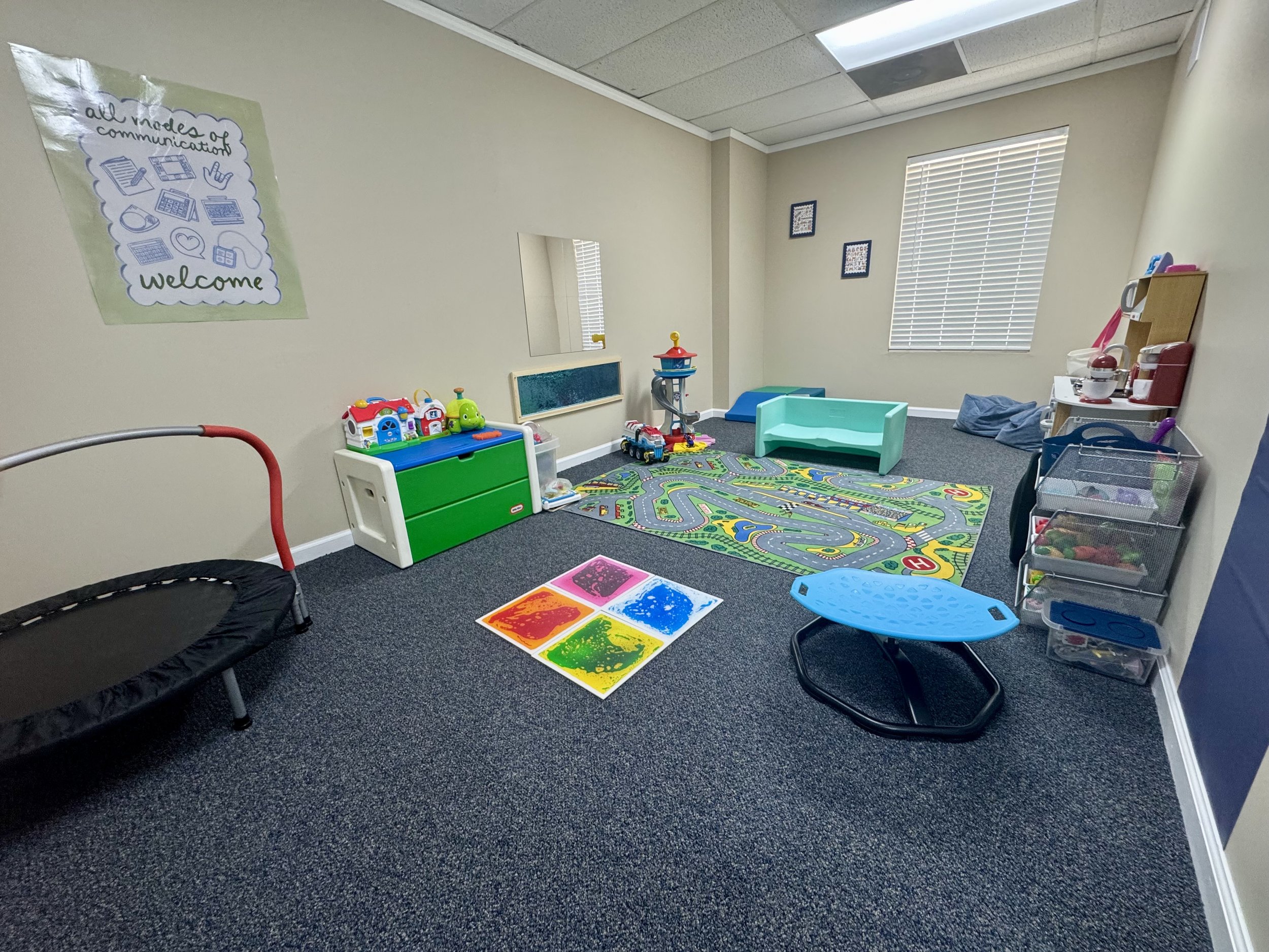 A playroom with a colorful city-themed rug, a trampoline, toy storage bins, and a small kitchen set against beige walls with window blinds, a mirror, and framed pictures.