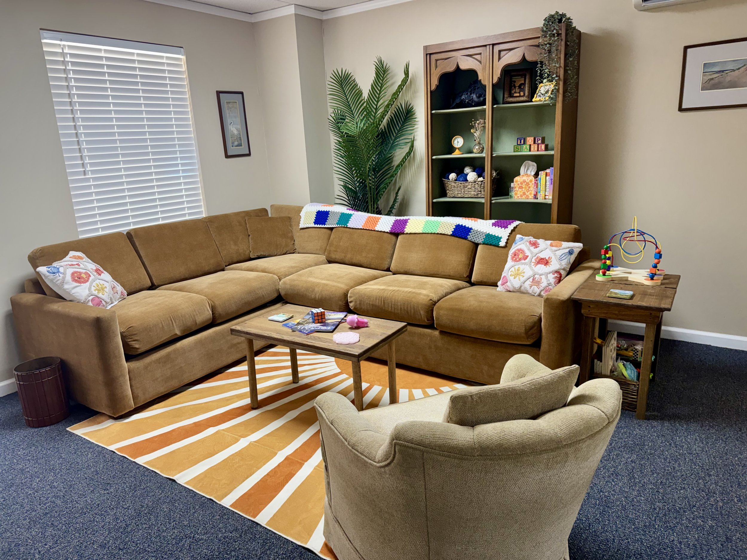 Living room with a large brown sectional sofa, a wooden coffee table with toys and magazines, a beige armchair, a striped orange and white rug, a tall green plant, a wooden bookshelf with decor and books, white blinds on the window, and framed pictures on the walls.