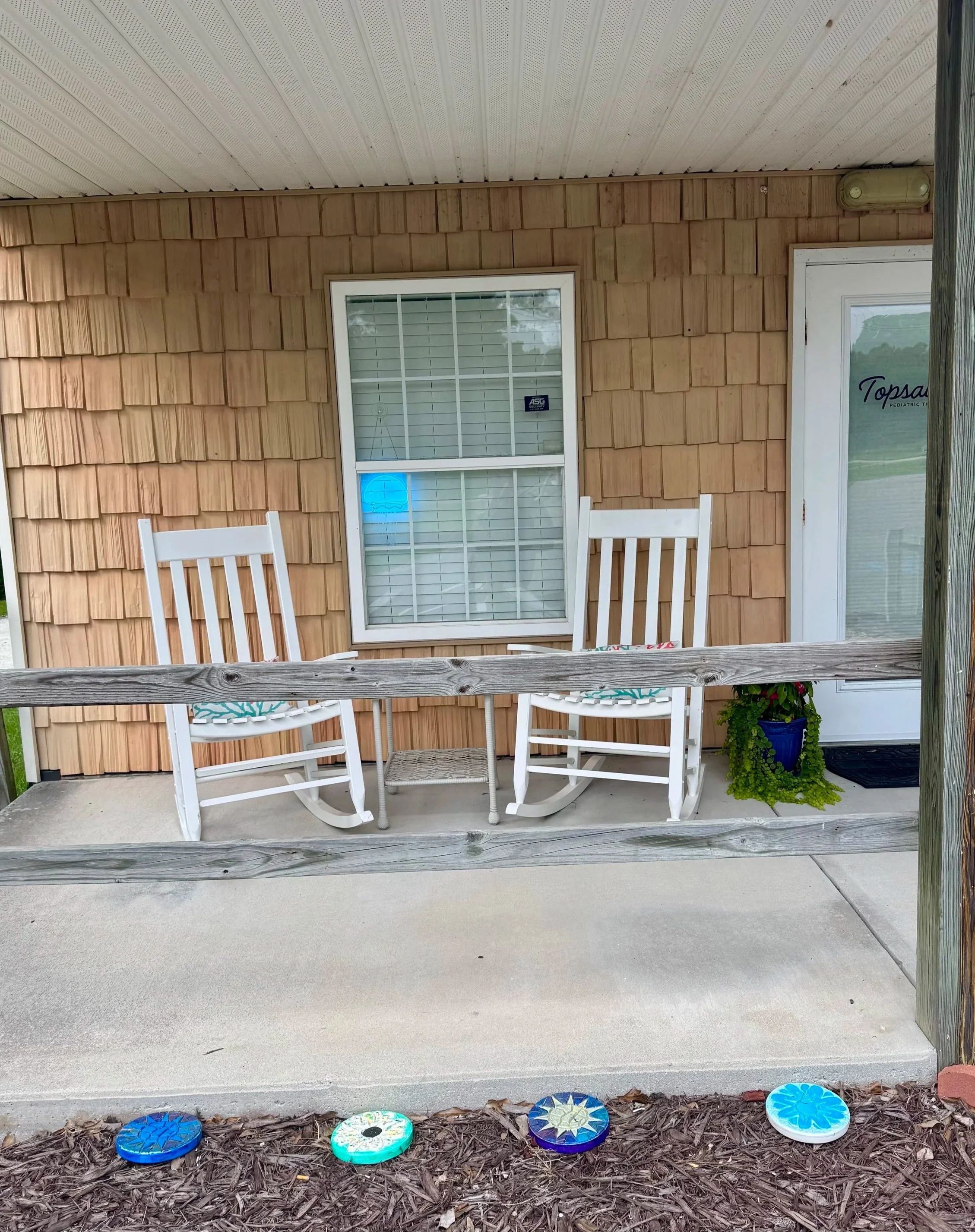 On a porch, there are four white chairs with a table covered with a cloth, in front of a house with tan wooden siding. A small plant in a blue pot is on the right side near the door, which has a sign saying 'Topsail Pediatric T...' The porch has a wooden railing, and beyond it, there is a mulched garden area with several decorated ceramic stepping stones.