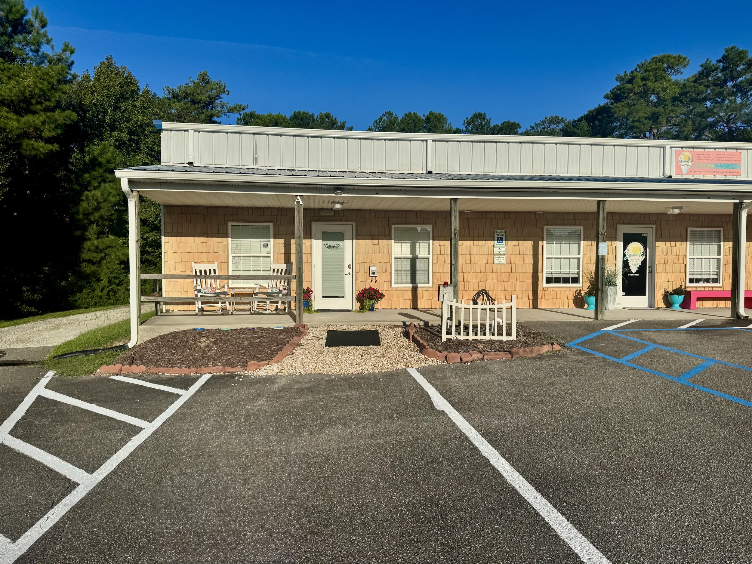 Front view of a single-story building with a covered porch, surrounded by trees, with parking spaces in front and a clear blue sky.