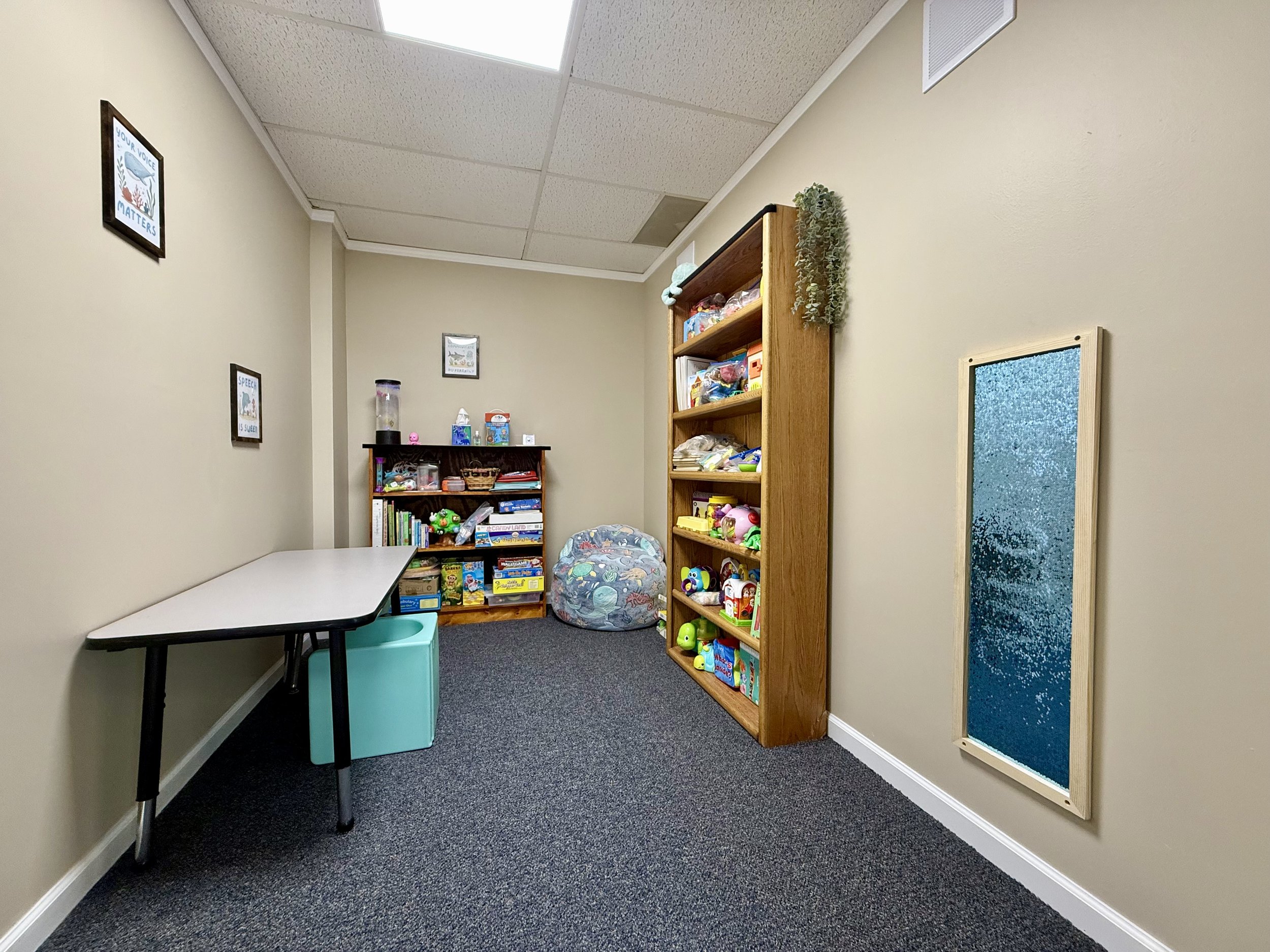 A small room with beige walls, a white ceiling with fluorescent lights, and a dark gray carpet. There is a long white table on the left with a teal storage unit underneath. In the corner, there are two wooden bookshelves filled with toys, books, and art supplies. The right wall has a corkboard and a small blue window.