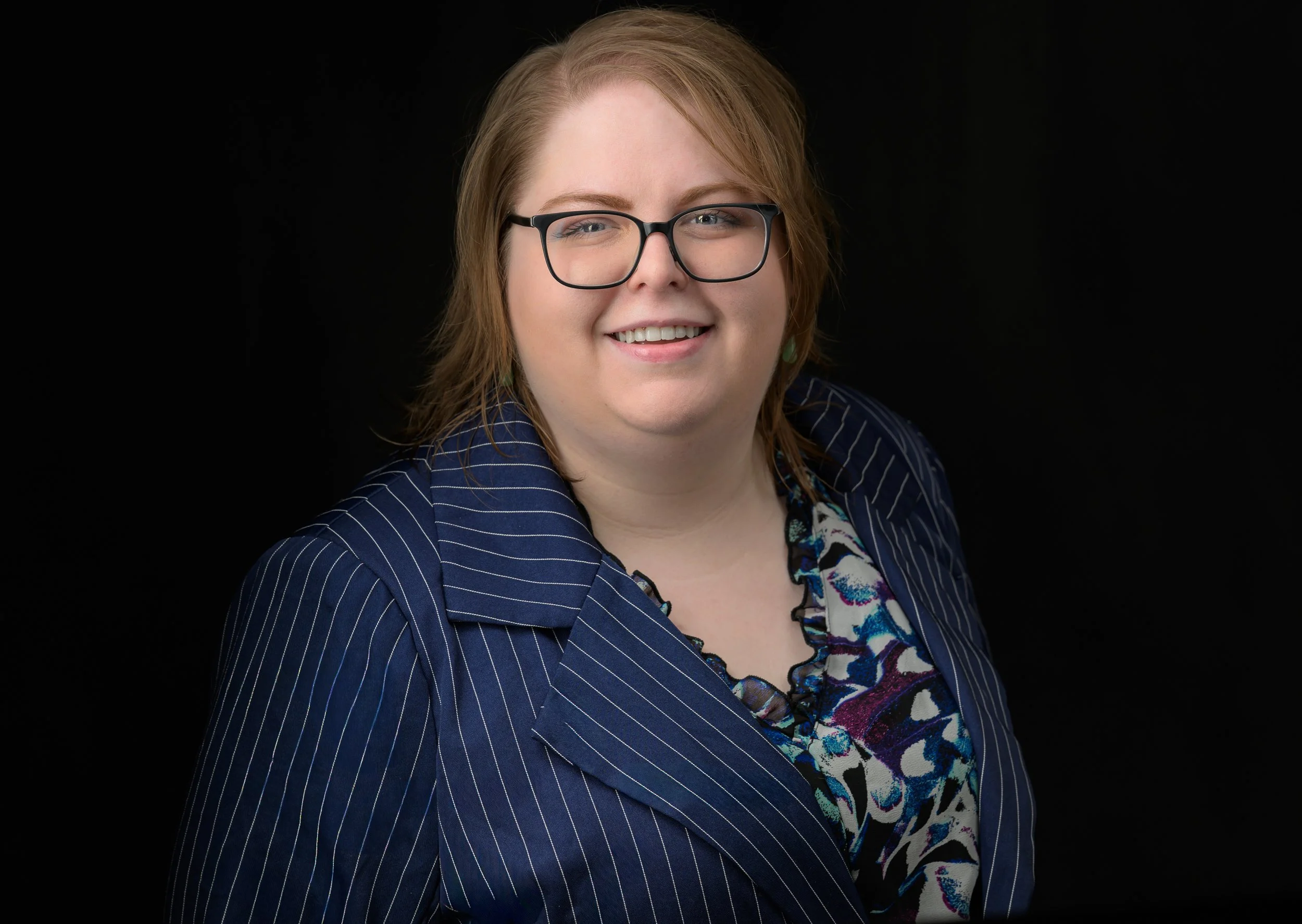 Young white woman professional smiling wearing glasses blouse and blazer on black background.