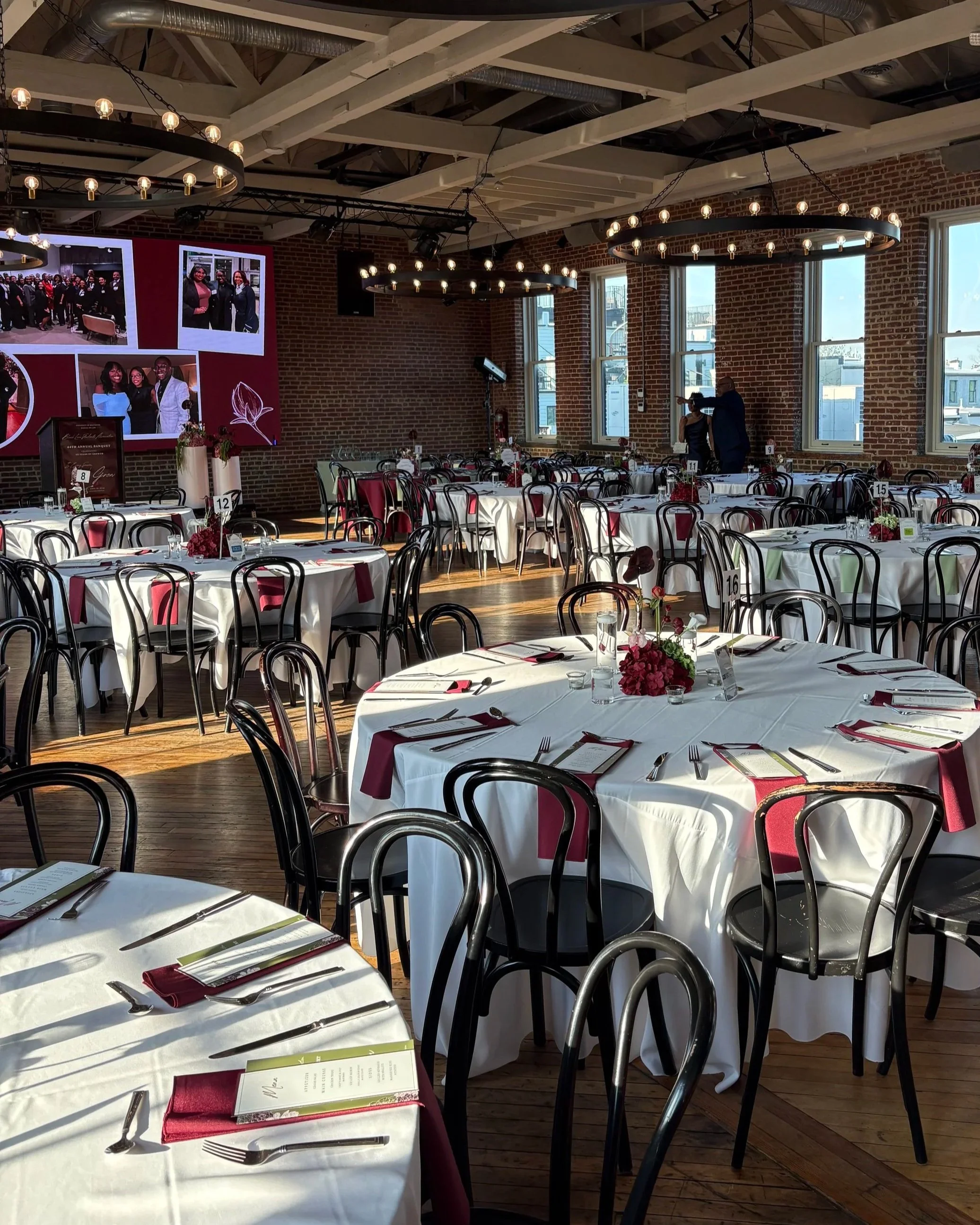 Elegant event styling and banquet tablescape with white linens, burgundy napkins, floral centerpieces, custom menu cards, and black bentwood chairs in an industrial-style Baltimore, MD event venue.