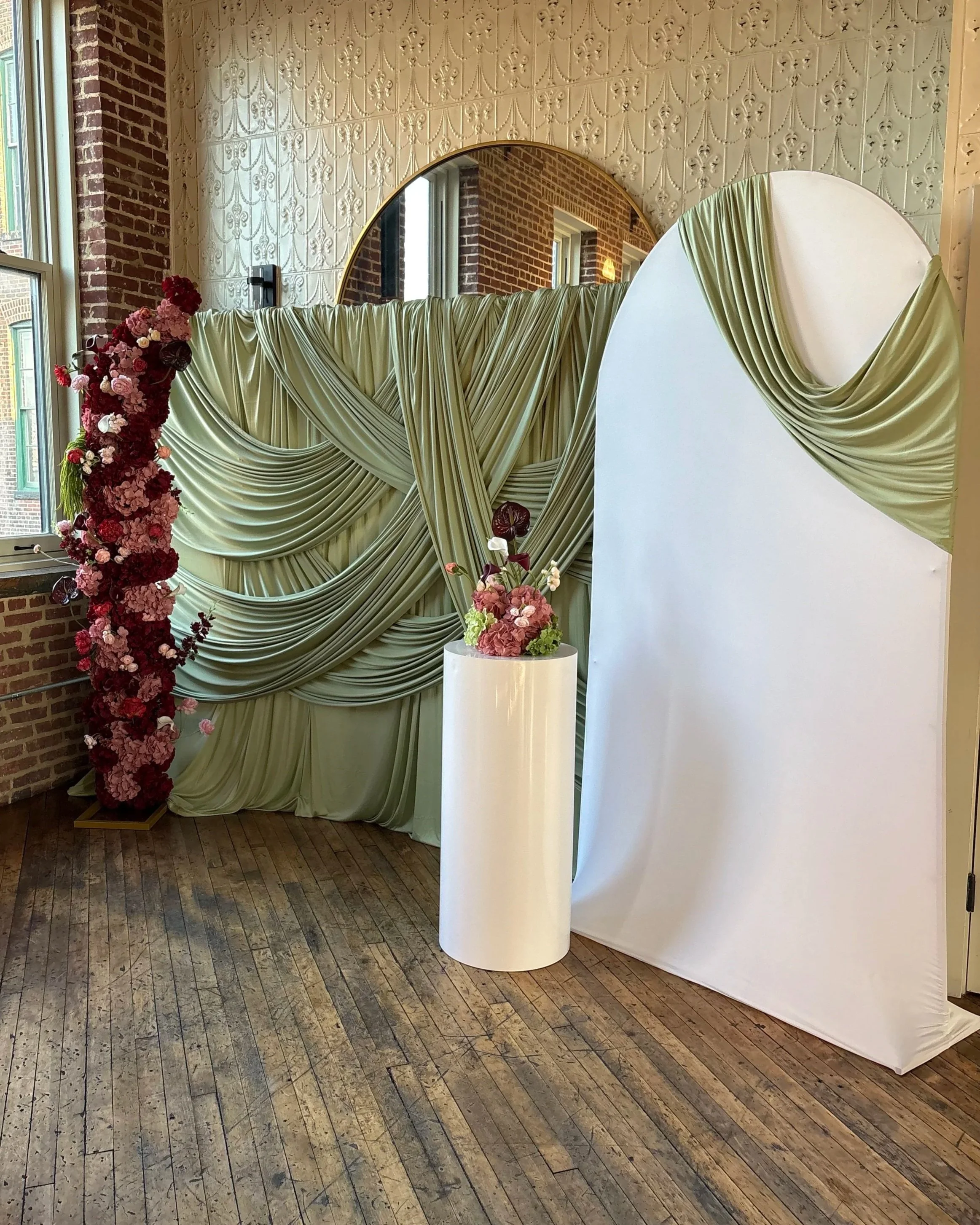 Elegant event backdrop with neutral draping, a tall pink and red floral arrangement, white pedestal, and modern mirror accent.