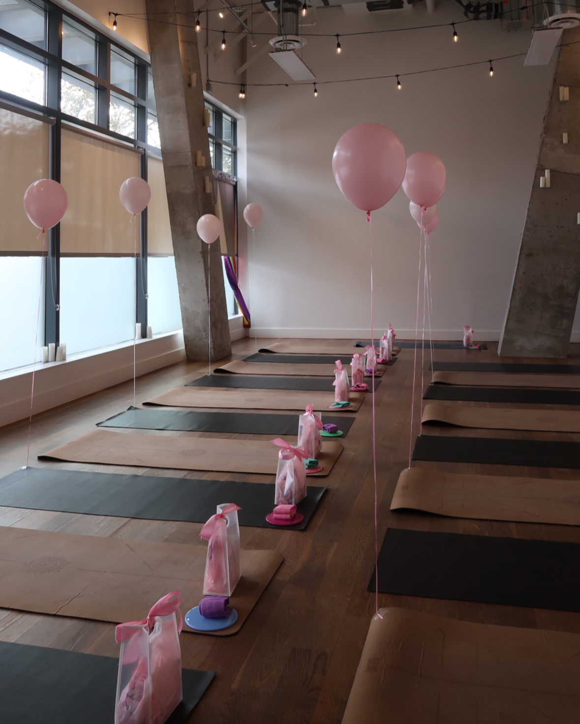 Yoga mats arranged in a row inside a studio decorated with pink balloons and wrapped gift bags at each mat, possibly set up for a celebratory or themed yoga class.