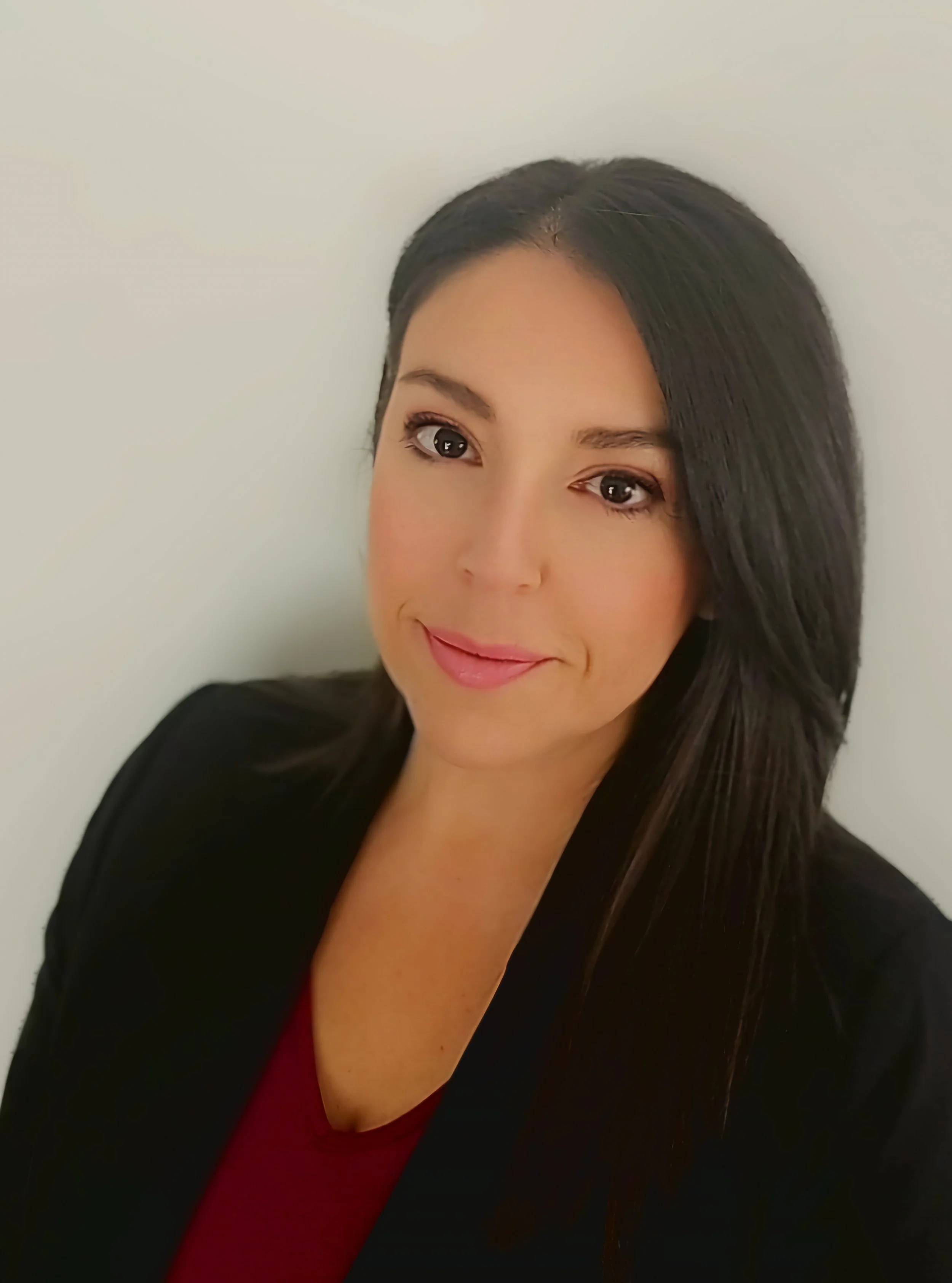 A woman with long dark hair, wearing a black blazer and a red top, smiling slightly in front of a plain white wall.