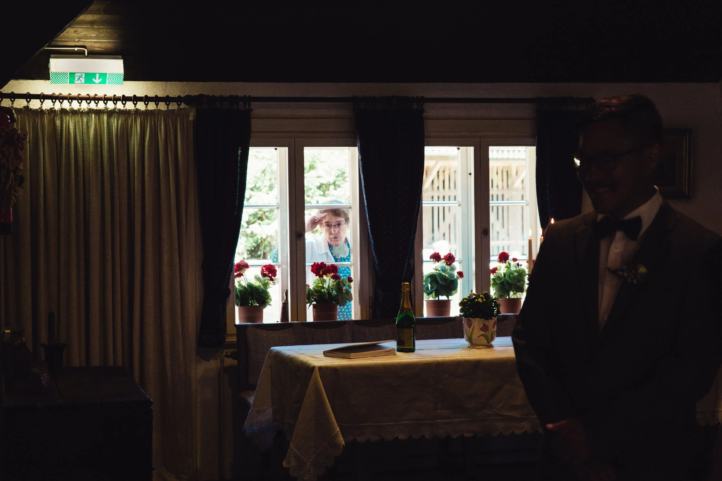 Ein Mann im Anzug und mit Brille steht im Schatten eines Raums, während eine Frau durch ein Fenster im Hintergrund einen Blick hineinwirft. Auf dem Tisch im Raum stehen eine Flasche Sekt, eine Zeitschrift und eine Topfpflanze.