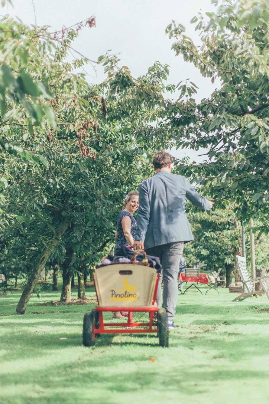Ein Mann und eine Frau in einem Park mit grünen Bäumen und Rasen, die miteinander lachen und spazieren gehen. Im Vordergrund befindet sich ein rotes Kinderrad mit einem Beutel, auf dem „Pinolino“ steht.