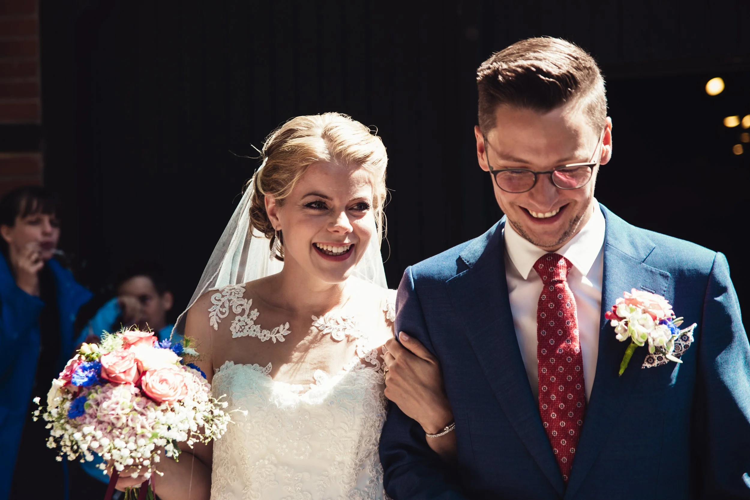 Ein glückliches Brautpaar bei ihrer Hochzeit, die Braut trägt ein weißes Kleid und hält einen Blumenstrauß, der Bräutigam trägt einen blauen Anzug mit roter Krawatte und Brille, sie lachen zusammen.