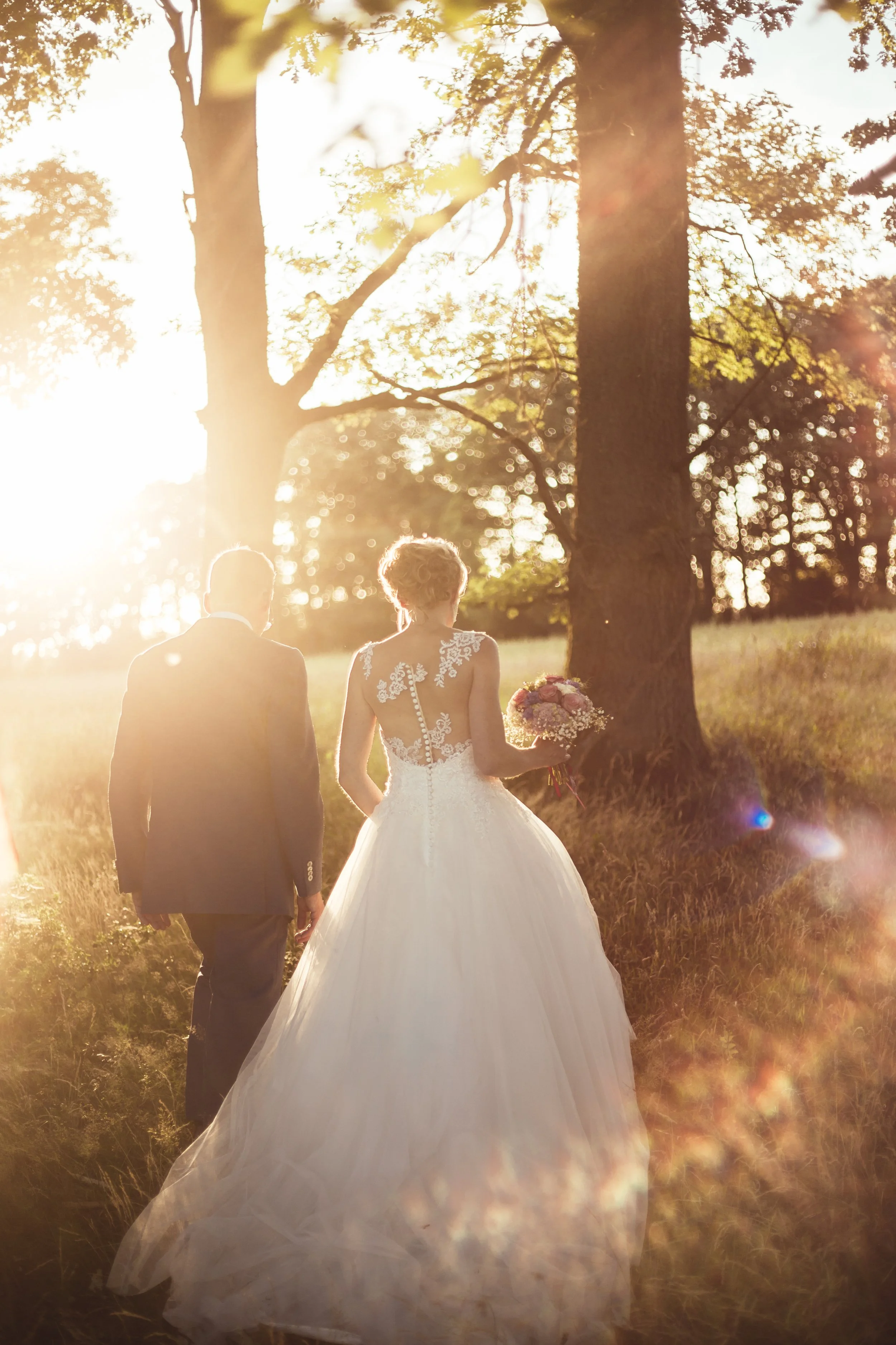 Ein Brautpaar wandert bei Sonnenuntergang durch einen Wald, die Braut trägt ein weißes Hochzeitskleid und hält einen Blumenstrauß.