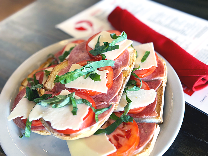 Open-faced sandwich with tomatoes, mozzarella, basil, salami, and cheese on toasted bread, served on a white plate with a red napkin and menu in background.