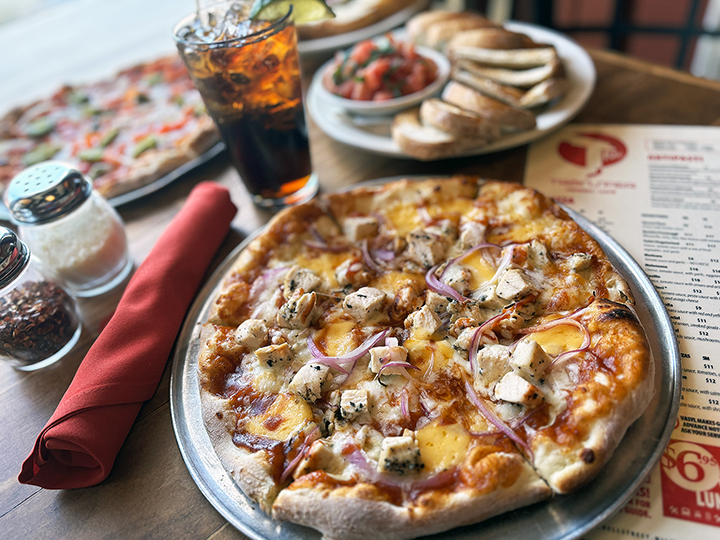 A pizza with chicken, onions, and cheese on a metal tray, accompanied by a glass of soda, a small bowl of grated cheese, salt and pepper shakers, a red napkin, and a side salad with pita bread on a restaurant table.