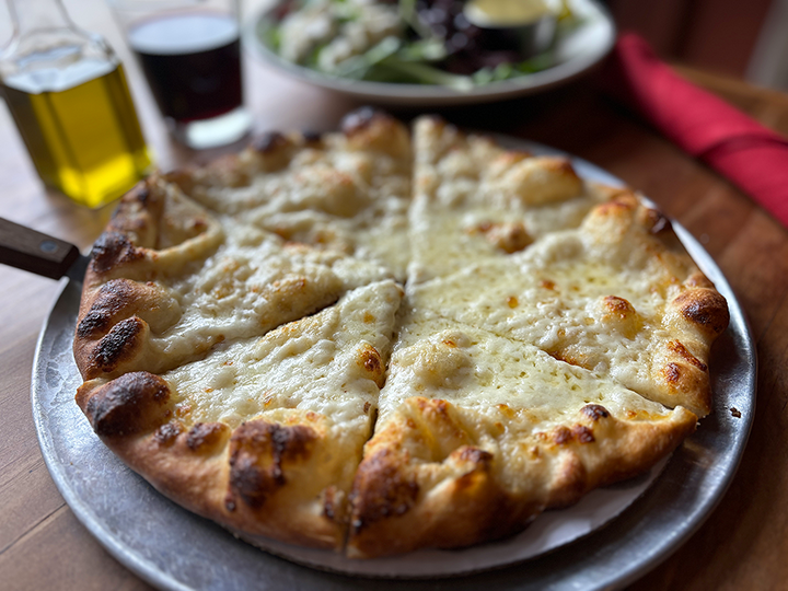 Cheese pizza on a metal tray in a restaurant setting with drinks in the background