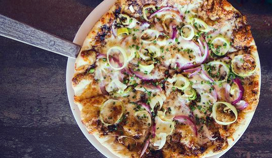 A whole pizza topped with sliced red onions, green onions, and cheese on a white plate, placed on a dark wooden surface.