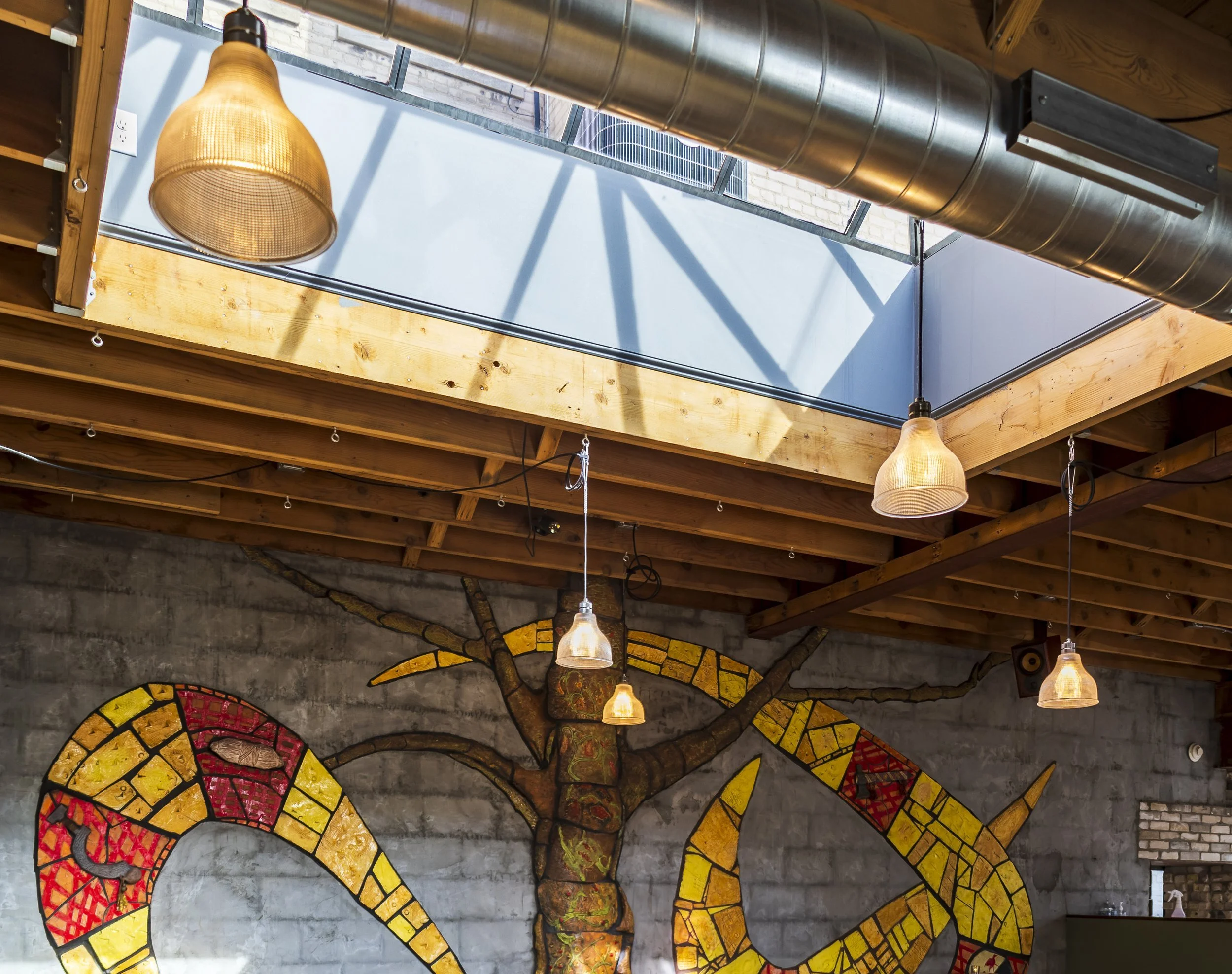 Interior of a restaurant with a wooden ceiling, hanging light fixtures, exposed ductwork, skylight, and a colorful dragon mural on a concrete wall.