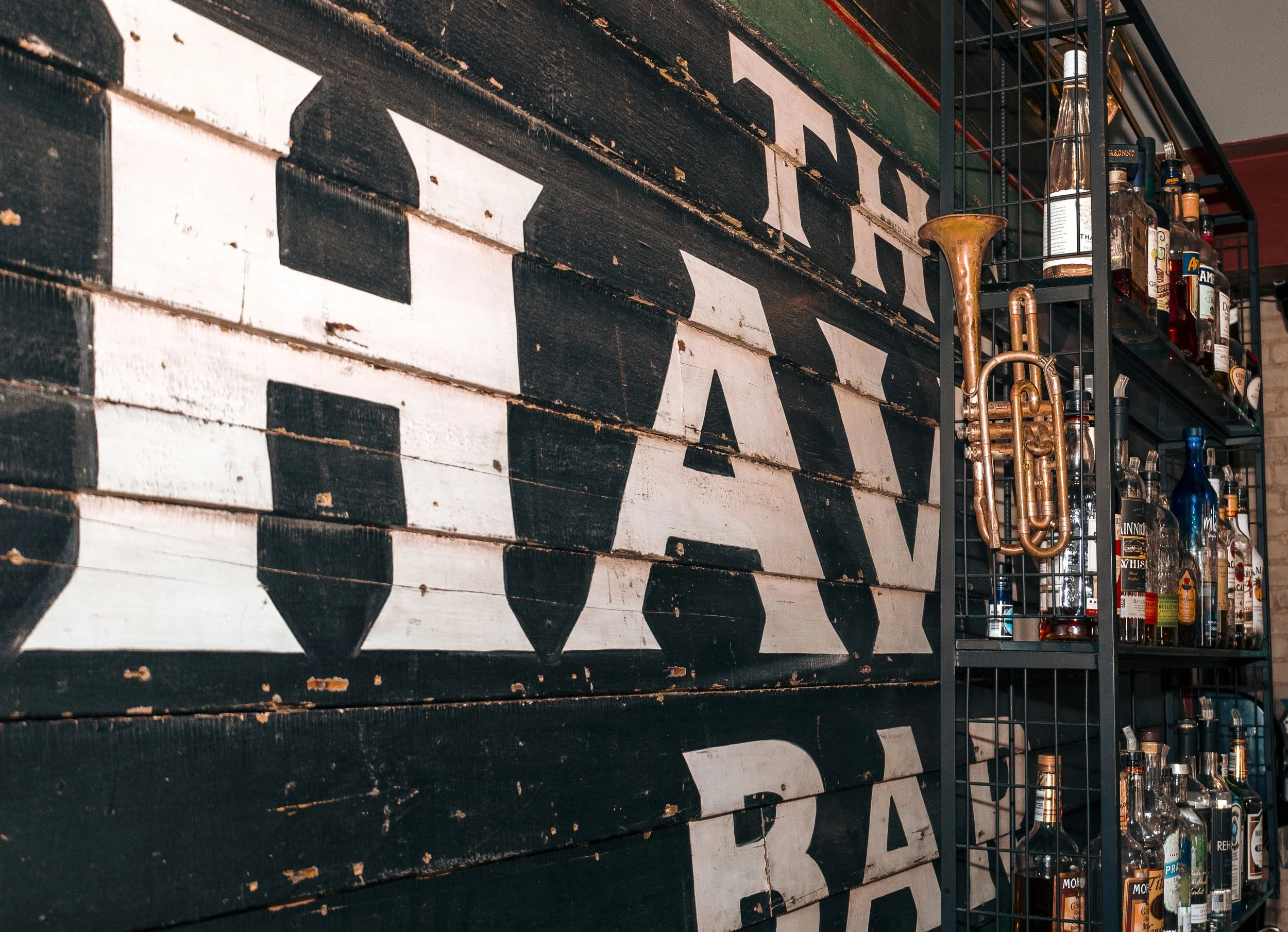 Close-up of a large vintage wooden sign with the words 'JACK DANIEL'S' painted in black and white, alongside a black metal shelf filled with liquor bottles and a copper instrument shaped like a trumpet or bugle.
