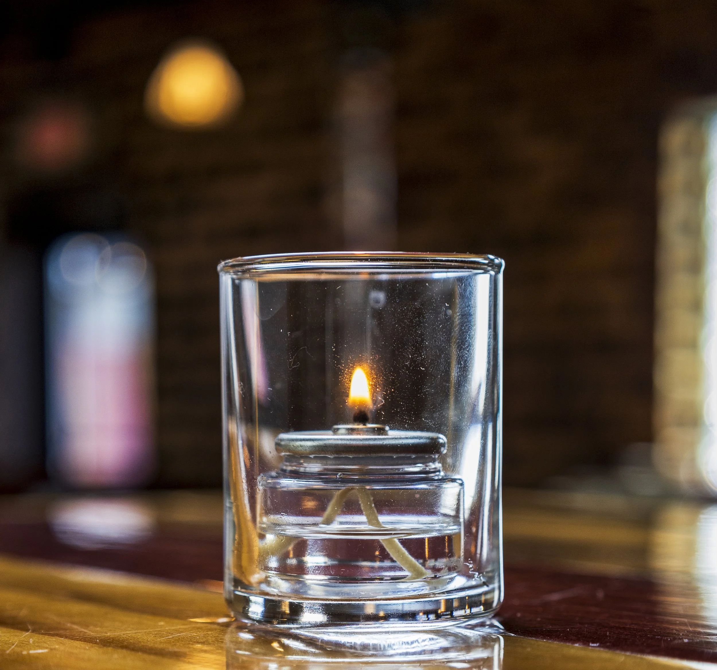Close-up of a floating candle in a glass container with visible wicks, lit with a small flame, on a wooden surface in a cozy indoor setting.