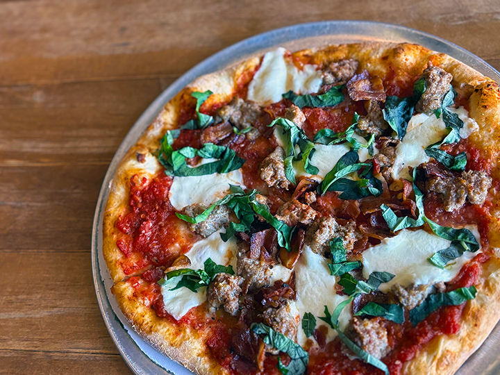 Close-up of a freshly baked pizza with sausage, bacon, mozzarella cheese, tomato sauce, and basil on a wooden table.