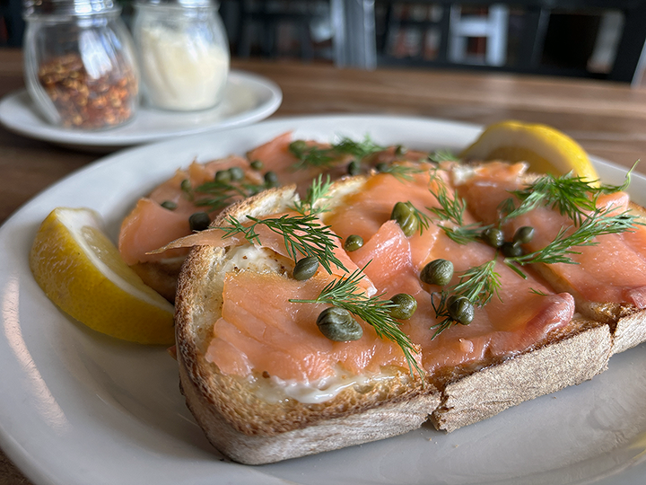 Close-up of smoked salmon on toasted bread with capers, fresh dill, and lemon wedges on a white plate.