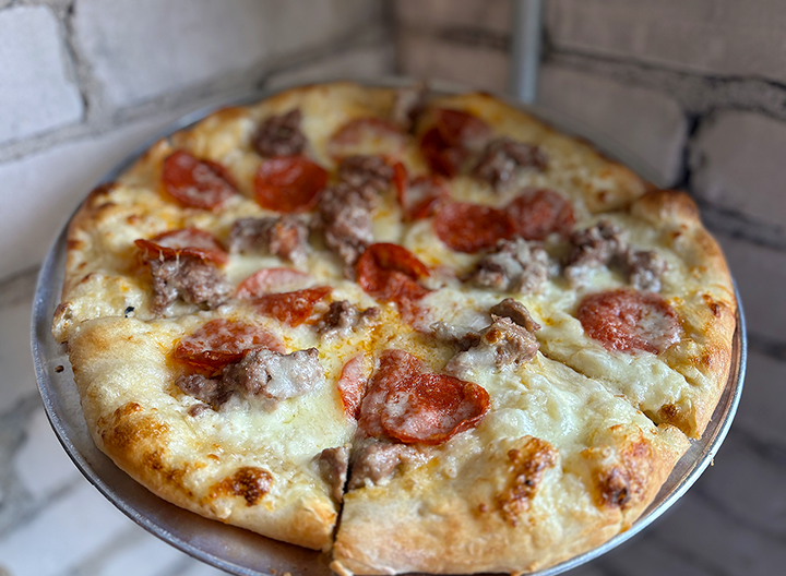 Pizza with pepperoni, sausage, and melted cheese on a metal tray against a brick wall background.