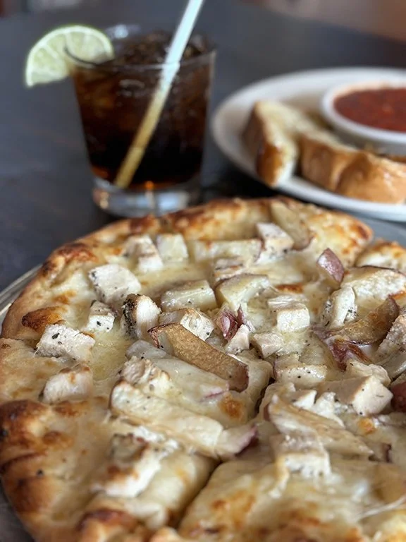 Close-up of a chicken pizza with cheese and chopped chicken pieces on a wooden table. In the background, there is a glass of soda with a lime wedge, a plate with breadsticks and marinara sauce, and part of a dipping plate.
