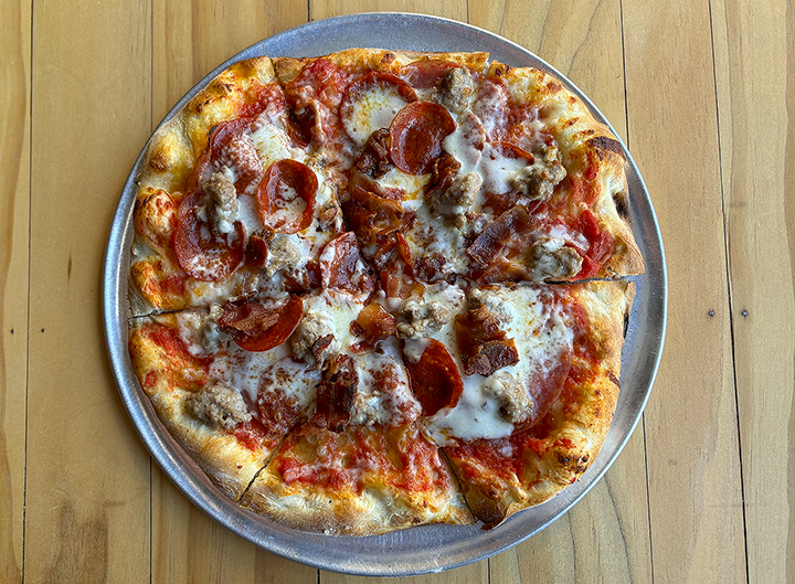 A pizza with pepperoni, sausage, and bacon slices on a metal tray, placed on a wooden table.