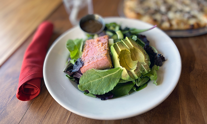 Salad with avocado slices, salmon slice, mixed greens, cherry tomatoes, and dressing on a white plate.