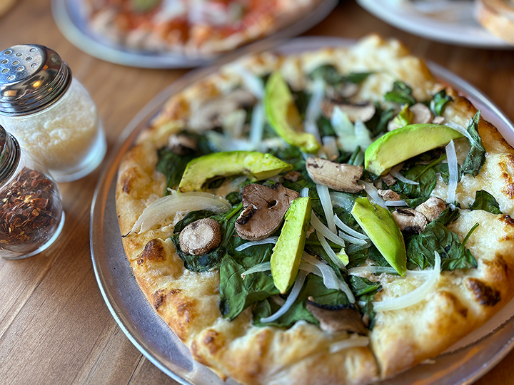 Vegetarian pizza with avocado slices, mushrooms, onions, and spinach on a wooden table, with salt and pepper shakers nearby.