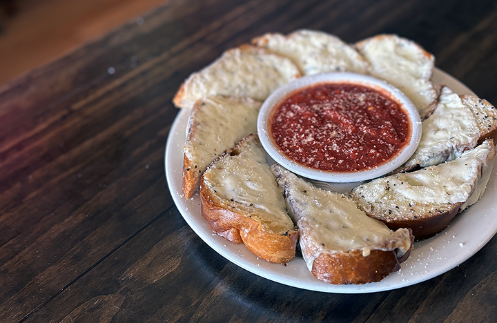 A white plate with slices of toasted bread topped with cheese, served with a small bowl of marinara sauce in the center, on a dark wooden table.