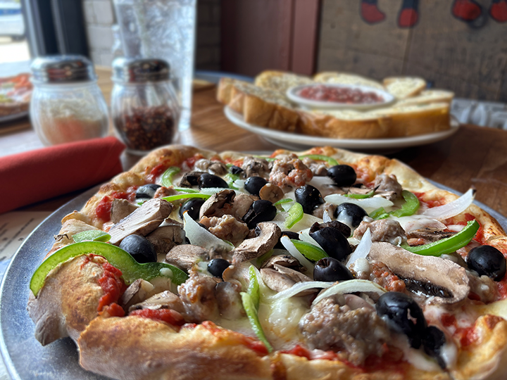 Close-up of a pizza topped with black olives, sliced mushrooms, green bell peppers, onions, and cheese. In the background, there is a plate with slices of bread and a dish of sauce, with condiment jars and a glass of water on the table.